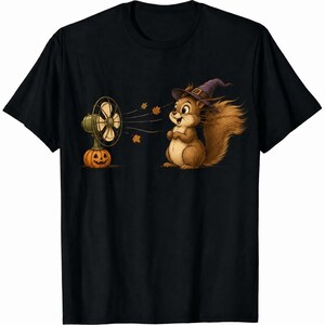 May include: A black short-sleeve t-shirt featuring a graphic of a brown squirrel wearing a purple witch hat. The squirrel stands near a vintage-style fan mounted on a carved jack-o'-lantern, with orange autumn leaves blowing in the air.