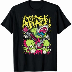 May include: A black short-sleeve t-shirt featuring a graphic print of a green, furry, shark-like creature playing a pink electric guitar. The text ATTACK ATTACK! appears in yellow above the creature, with blue and pink paint splatter accents.