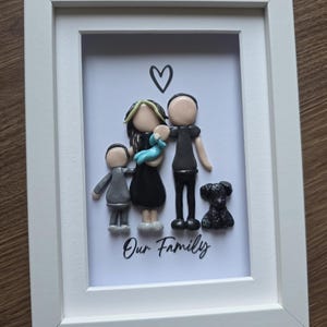 May include: A white framed wall art piece featuring a 3D clay sculpture of a family of four and a dog. The figures include two adults, one child, and a baby in blue, with a black heart icon above and the text Our Family printed in black script.