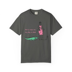 May include: Dark gray t-shirt with a humorous graphic. The design features a cartoon alligator, a woman in a pink dress, and the text "Don't rush me, my dog is dead." The shirt is a casual wear item.