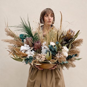 Puede incluir: Un arreglo grande y ancho de flores y follaje secos en un cuenco de madera. El ramo presenta proteas de color púrpura oscuro, rosas blancas, hierba de la pampa esponjosa, hojas de eucalipto verdes, motas de algodón y hierbas secas finas.