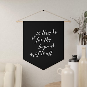 May include: A black fabric wall banner featuring the white text "to live for the hope of it all" in a cursive font, accented by small four-pointed star icons. The banner is suspended from a wooden dowel with a thin string for hanging.