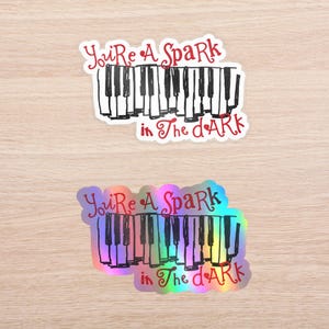 May include: Two stickers featuring a black and white piano keyboard illustration with the red text "You're A Spark in The dArk" written above and below the keys. One sticker has a white background, while the other has a holographic finish.