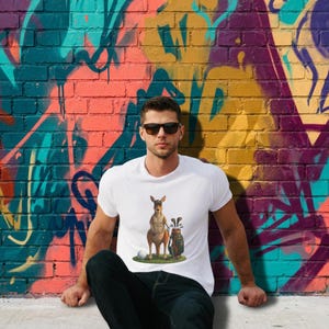 May include: A white short-sleeved t-shirt featuring a graphic of a brown kangaroo standing next to a golf bag filled with clubs and a white golf ball on the ground. The shirt is worn with dark blue jeans and black sunglasses.