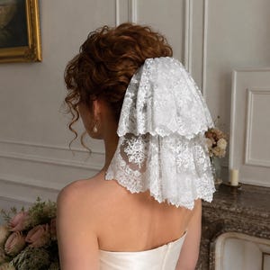 May include: A two-tiered white lace veil with intricate floral embroidery and scalloped edges, secured in the hair. The veil features a delicate, semi-sheer mesh base with detailed needlework patterns throughout the fabric, creating a layered effect.