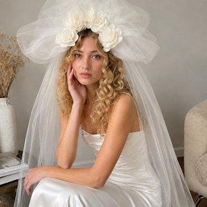 May include: A white bridal veil featuring a voluminous tulle headpiece adorned with a row of five fabric flowers. The sheer, layered tulle extends downward, creating a soft, draped effect. The accessory is paired with a simple white satin dress.