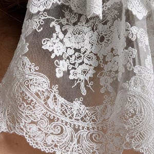 May include: A white lace fabric featuring intricate floral embroidery and detailed scalloped edges. The delicate mesh material displays a repeating pattern of blossoms and swirling botanical motifs, creating a textured and semi-sheer appearance.