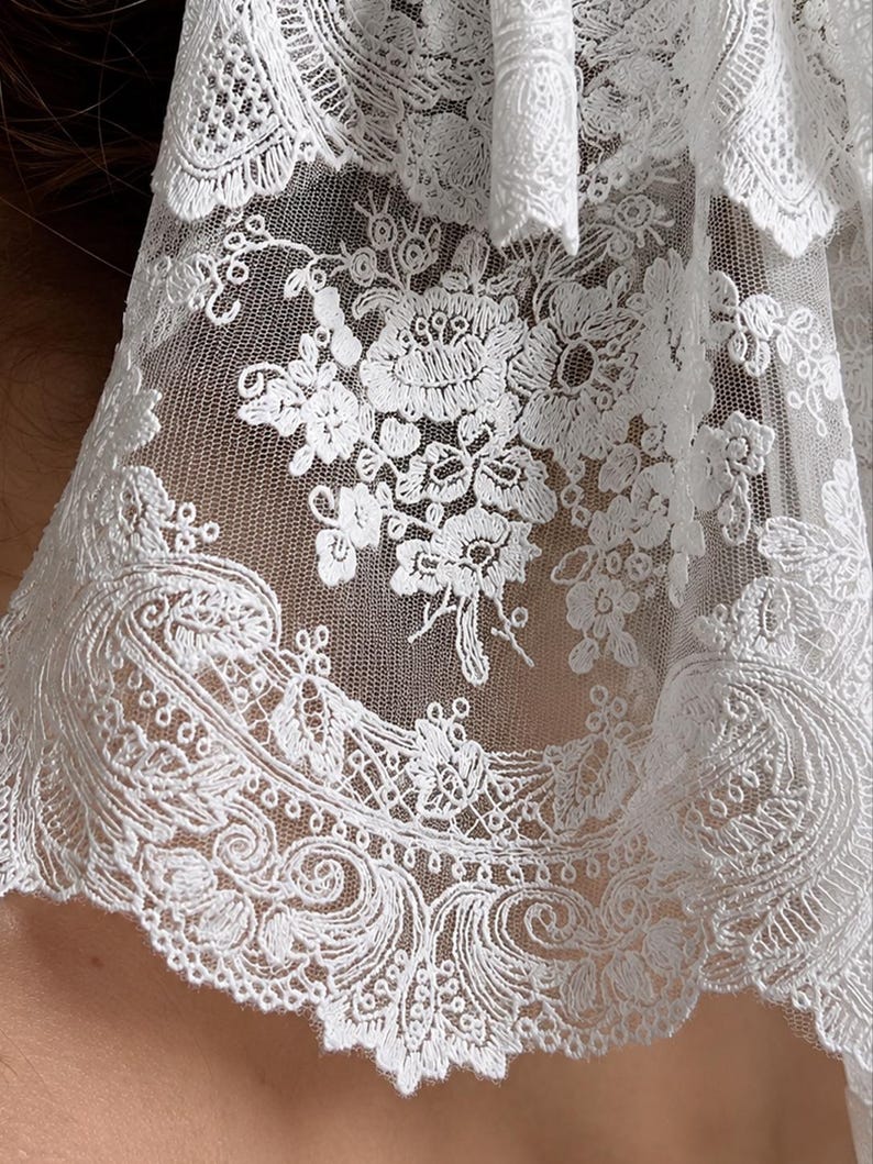 May include: A white lace fabric featuring intricate floral embroidery and detailed scalloped edges. The delicate mesh material displays a repeating pattern of blossoms and swirling botanical motifs, creating a textured and semi-sheer appearance.