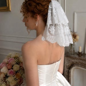 May include: A white two-tiered lace veil with intricate floral embroidery and scalloped edges, secured at the back of the head. The veil features a delicate, sheer mesh base with detailed needlework patterns throughout the fabric layers.