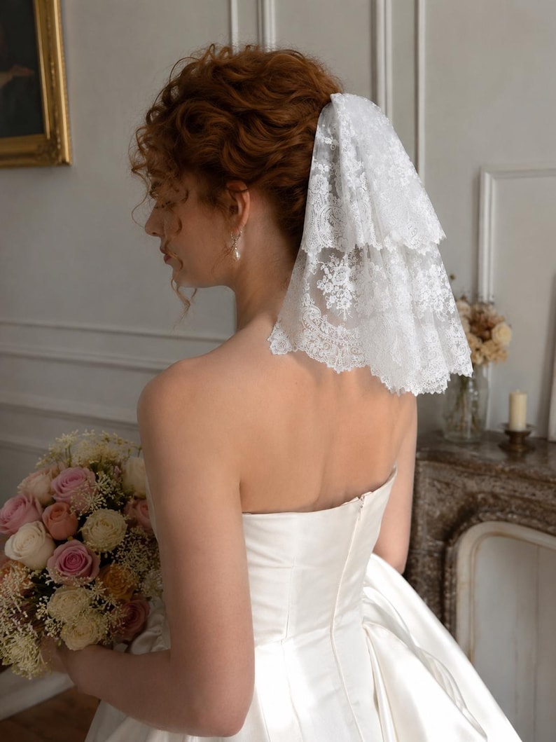 May include: A white two-tiered lace veil with intricate floral embroidery and scalloped edges, secured at the back of the head. The veil features a delicate, sheer mesh base with detailed needlework patterns throughout the fabric layers.