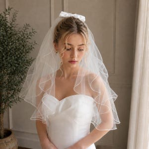 May include: A white bridal veil featuring two tiers of sheer tulle with a delicate rolled edge finish. The veil is secured at the top with a matching white satin bow, creating a soft, layered effect that drapes gracefully over the shoulders.
