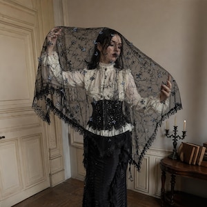 May include: A black lace veil featuring delicate blue floral embroidery and a scalloped edge, worn over a cream-coloured long-sleeved blouse with a high ruffled collar, a black structured corset, and a long black skirt with layered fabric details.