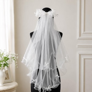 May include: A white two-tier bridal veil featuring a sheer tulle fabric with a scalloped edge. The veil is adorned with small white ribbon bows placed along the hem and at the top, where a large, structured bow secures the layers together.