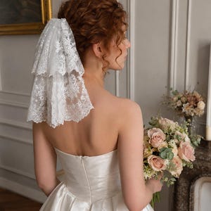 May include: A two-tiered white lace veil with intricate floral embroidery and scalloped edges, secured at the back of the head. The veil features a delicate, sheer mesh base with detailed needlework patterns throughout the fabric layers.