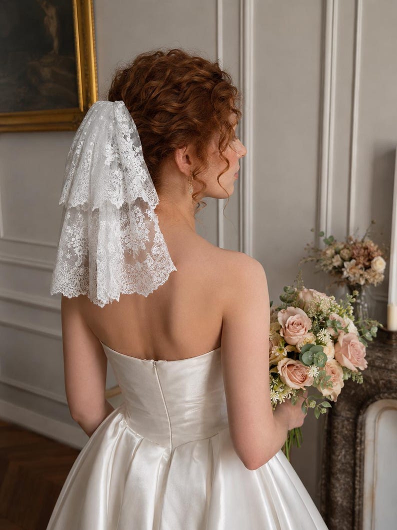 May include: A two-tiered white lace veil with intricate floral embroidery and scalloped edges, secured at the back of the head. The veil features a delicate, sheer mesh base with detailed needlework patterns throughout the fabric layers.