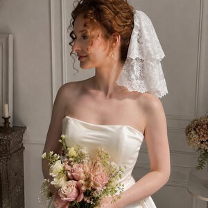 May include: A white two-tiered lace veil with scalloped edges worn in the hair, paired with a strapless ivory satin gown and a bouquet of pink and white roses, greenery, and small white filler flowers held in the hands.