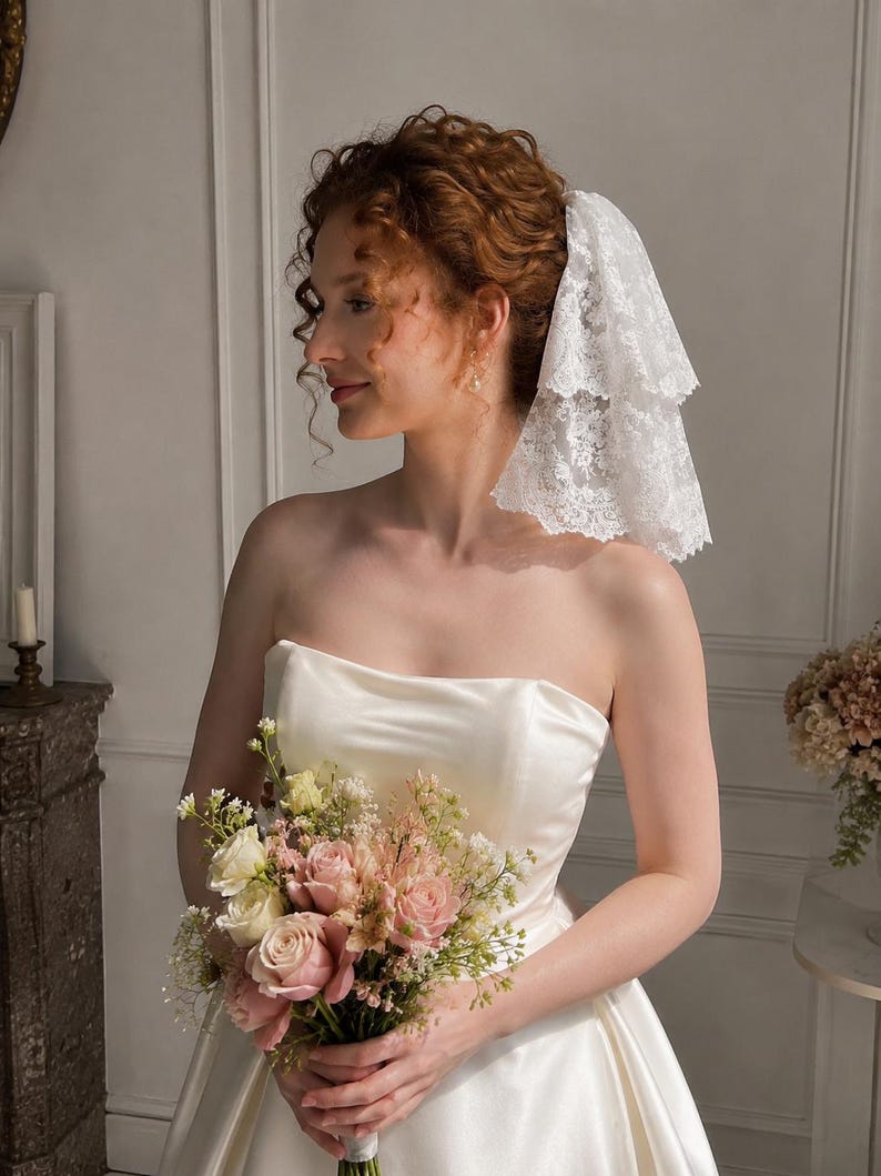 May include: A white two-tiered lace veil with scalloped edges worn in the hair, paired with a strapless ivory satin gown and a bouquet of pink and white roses, greenery, and small white filler flowers held in the hands.