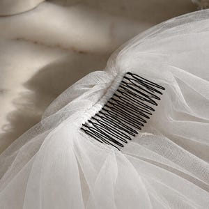 May include: A white tulle veil attached to a black metal hair comb. The fine, sheer netting is gathered and securely fastened along the top edge of the comb, creating soft, layered folds that drape downwards from the dark, multi-pronged fastener.
