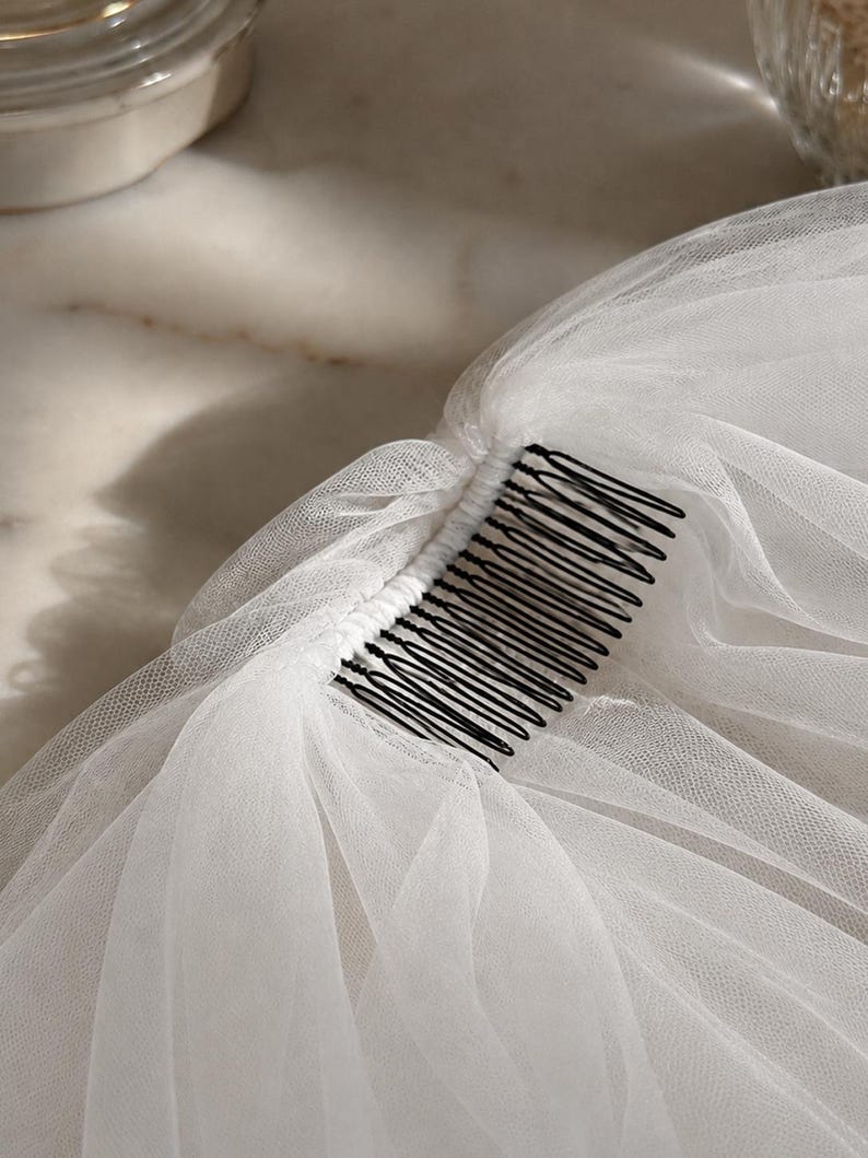May include: A white tulle veil attached to a black metal hair comb. The fine, sheer netting is gathered and securely fastened along the top edge of the comb, creating soft, layered folds that drape downwards from the dark, multi-pronged fastener.