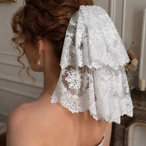 May include: A two-tiered white lace veil featuring intricate floral embroidery and scalloped edges. The sheer fabric is layered to create volume, with detailed patterns visible throughout the lace. The veil is attached to the hair, cascading down.