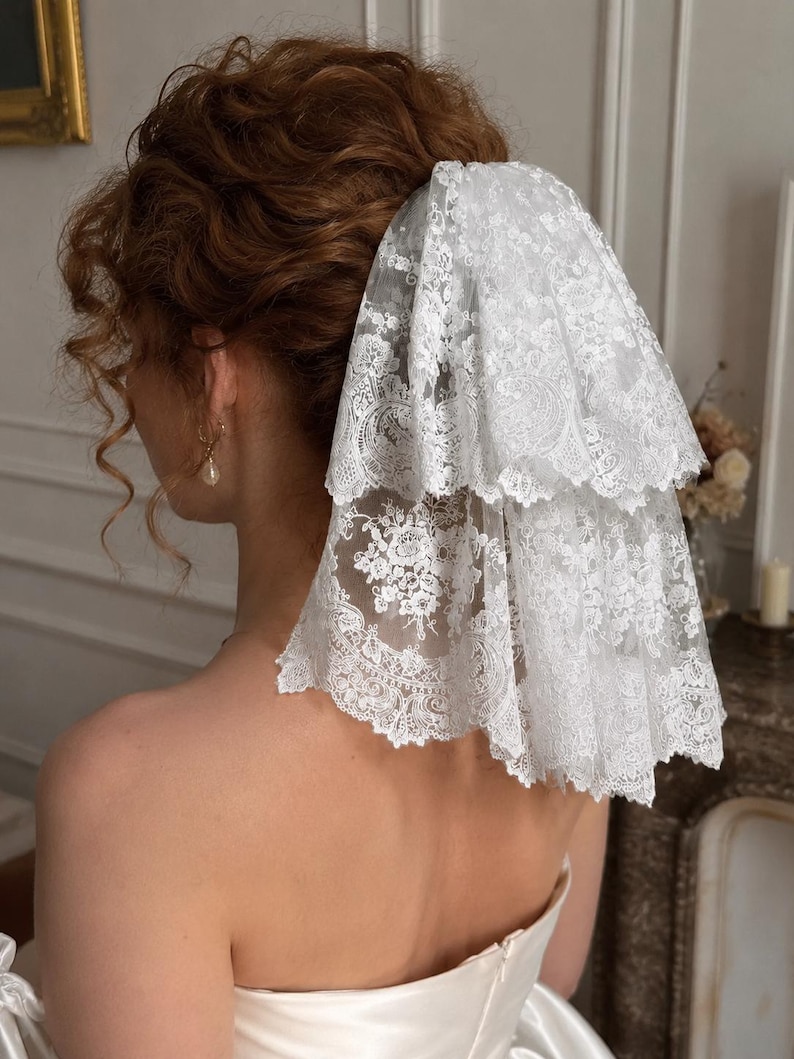 May include: A two-tiered white lace veil featuring intricate floral embroidery and scalloped edges. The sheer fabric is layered to create volume, with detailed patterns visible throughout the lace. The veil is attached to the hair, cascading down.