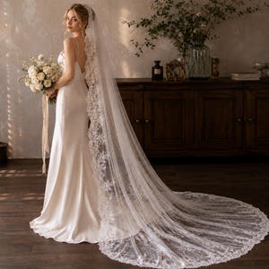 May include: A floor-length white wedding dress with thin straps and a low back, paired with a long, sheer cathedral-length veil featuring intricate white floral lace trim along the edges. The ensemble is completed by a bouquet of white flowers.