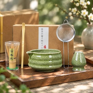 May include: A matcha tea set including a bamboo whisk in a clear case, a bamboo scoop, a green ceramic bowl with a spout, a metal mesh strainer, and a green ceramic whisk holder, all arranged in front of a cardboard box with Japanese text.