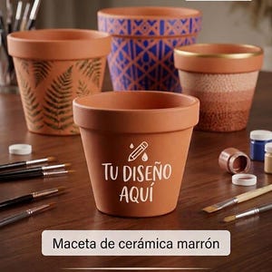 Può includere: Quattro vasi da fiori in terracotta con un diametro di 27 cm. Un vaso presenta la scritta bianca TU DISEÑO AQUÍ con un'icona a matita. Gli altri mostrano motivi a felce, disegni geometrici blu e un bordo dorato. Pennelli sono vicini.