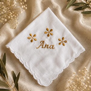 Puede incluir: Pañuelo blanco con borde festoneado, bordado con el nombre "Ana" en hilo dorado y tres diseños florales dorados. El pañuelo se muestra sobre un fondo de tela beige con detalles florales.