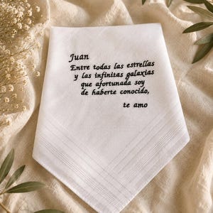 Puede incluir: Un pañuelo blanco con texto bordado en negro que dice: Juan, Entre todas las estrellas y las infinitas galaxias que afortunada soy de haberte conocido, te amo. La tela presenta un detalle de borde con rayas sutiles en la parte inferior.