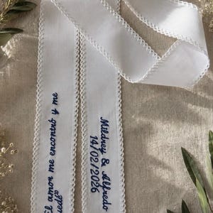 Könnte beinhalten: Ein langes weißes Stoffband mit gehäkelten Bogenkanten und dunkelblauer Stickerei mit dem Text "El amor me encontró y me quedé" sowie "Mildrey & Alfredo 14/02/2026". Das Band ist gefaltet und weist eine glatte, gewebte Textur auf.