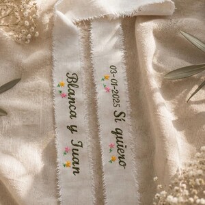 May include: Two off-white fabric ribbons with frayed edges featuring dark green embroidered text and small colorful floral accents. One ribbon reads Blanca y Juan, and the other reads 03-01-2025 Sí quiero, both in a cursive script font.