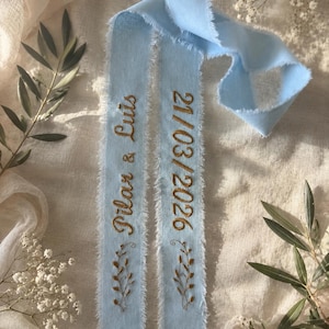Könnte beinhalten: Ein hellblaues Stoffband mit ausgefransten Kanten und goldener Stickerei, die auf einer Seite Pilar & Luis und auf der anderen 21/03/2026 zeigt, ergänzt durch kleine gestickte Blattzweige am unteren Ende jedes Bandabschnitts.