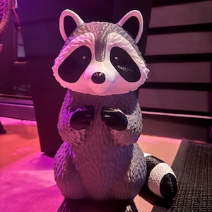 May include: A raccoon figurine featuring a gray and white textured body with black eye patches, a black nose, and black paws. The raccoon is in a standing position with its front paws held together near its chest and a striped tail extending to the side.