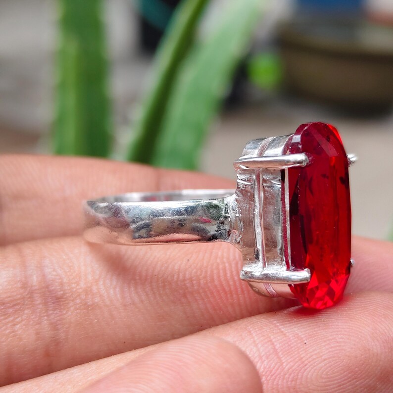 May include: A silver ring featuring a large, oval-cut, vibrant red gemstone held in place by a four-prong setting. The metal band has a smooth, polished finish, and the faceted red stone is positioned vertically, showcasing its deep crimson color.