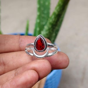 May include: A silver-toned ring featuring a central teardrop-shaped red gemstone. The band is split into two sections, with the lower portion forming a crescent moon shape that cradles the base of the teardrop stone. The metal has a polished finish.