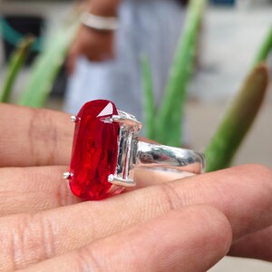 May include: A silver ring featuring a large, rectangular, faceted red gemstone held in place by four metal prongs. The band of the ring is smooth and polished, showcasing a metallic finish that contrasts with the vibrant, deep red color of the stone.