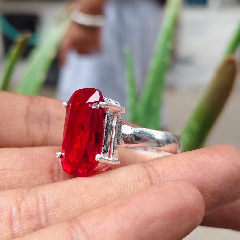 May include: A silver ring featuring a large, rectangular, faceted red gemstone held in place by four metal prongs. The band of the ring is smooth and polished, showcasing a metallic finish that contrasts with the vibrant, deep red color of the stone.