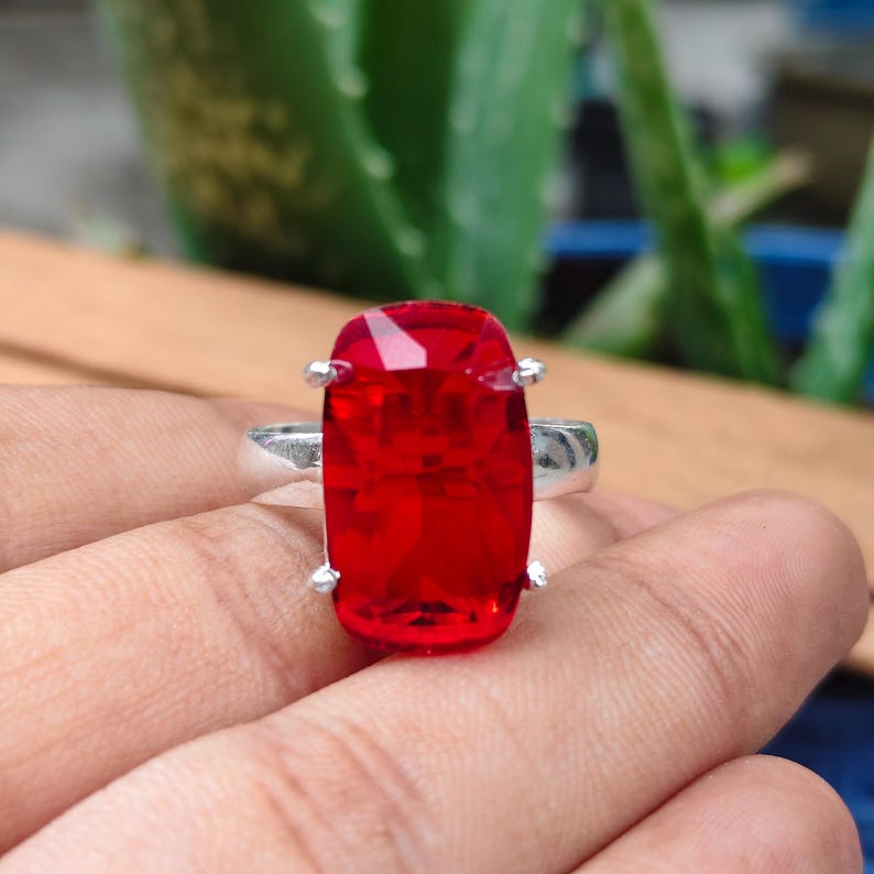 May include: A silver ring featuring a large, rectangular, faceted red gemstone set in a four-prong mounting. The stone has a vibrant, translucent appearance with sharp edges and a polished finish, held securely by a simple metallic band.
