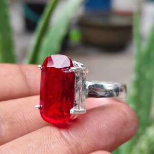 May include: A silver ring featuring a large, rectangular, faceted red gemstone held in place by four metal prongs. The band of the ring is smooth and metallic, showcasing a polished finish that reflects light across its surface.