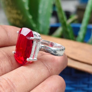 May include: A silver ring featuring a large, rectangular, faceted red gemstone held in place by four metal prongs. The band is smooth and metallic, showcasing a polished finish that reflects light across its surface and the vibrant red stone.