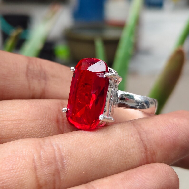 May include: A silver ring featuring a large, rectangular, faceted red gemstone held in place by four metal prongs. The band is smooth and metallic, showcasing a polished finish that reflects light across the surface of the vibrant red stone.