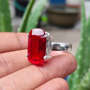 May include: A silver ring featuring a large, rectangular, faceted red gemstone held in place by four metal prongs. The stone has a vibrant, translucent appearance with sharp edges and a polished finish, set on a simple, smooth, metallic band.