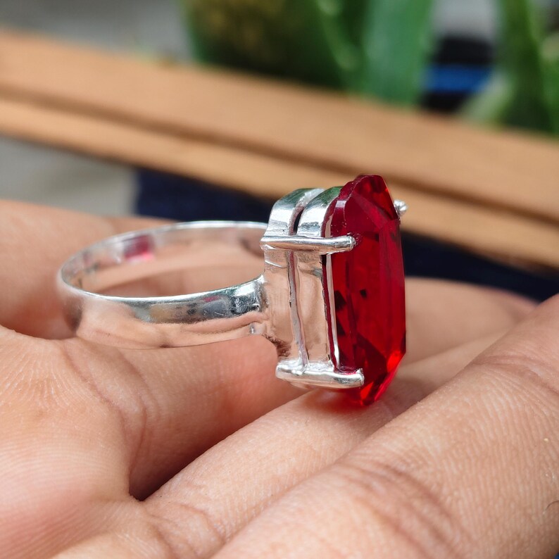 May include: A silver ring featuring a large, rectangular, faceted red gemstone set in a prong setting. The band is smooth and polished, connecting to a metallic frame that secures the vibrant red stone, which displays clear geometric facets.