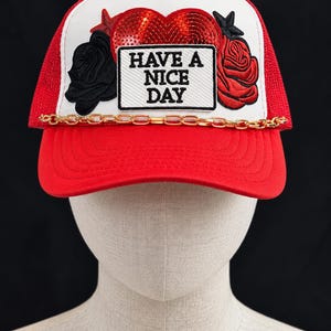 May include: A red and white trucker hat featuring a sequined red heart, a black rose, a red rose, and a rectangular patch with the text HAVE A NICE DAY. A gold-toned metal chain is attached across the base of the front panel above the red brim.