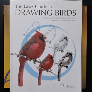 May include: A book titled "The Laws Guide to Drawing Birds" featuring illustrations of red cardinals perched on branches. The cover art includes sketches and the Audubon logo, with text by John Muir Laws.