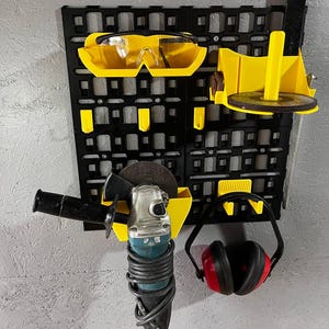 Könnte beinhalten: Ein schwarzer Wandorganizer mit gelber Schutzbrille, einem Schleifer, Gehörschutz und einer Scheibe. Der Organizer hat ein Gittermuster mit verschiedenen Schlitzen und Haltern für Werkzeuge und Zubehör. Der Schleifer ist silber und schwarz.
