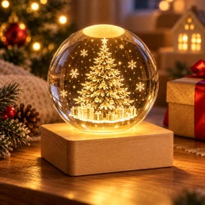 May include: A spherical glass ornament featuring an illuminated Christmas tree and wrapped gift boxes inside, mounted on a rectangular light-colored wooden base. The interior scene is accented with glowing star and snowflake patterns.