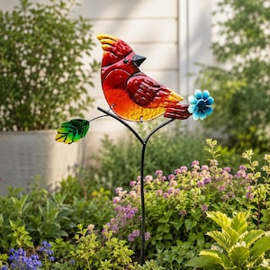 May include: A garden stake featuring a red and orange glass cardinal bird perched on a black metal branch. The bird has a yellow-tipped crest and a black beak. A green glass leaf is on the left side of the branch, and a blue metal flower is on the right.