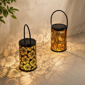 May include: Two black metal lanterns with handles. One lantern has a sunflower pattern, the other has a leaf design. Both lanterns cast floral and leaf-shaped shadows on the surface. The lanterns are illuminated with a warm, golden light.